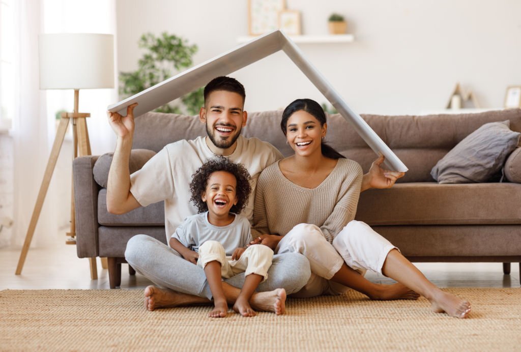 Mother, father, and child under a small roof on the living room floor, smiling at the camera